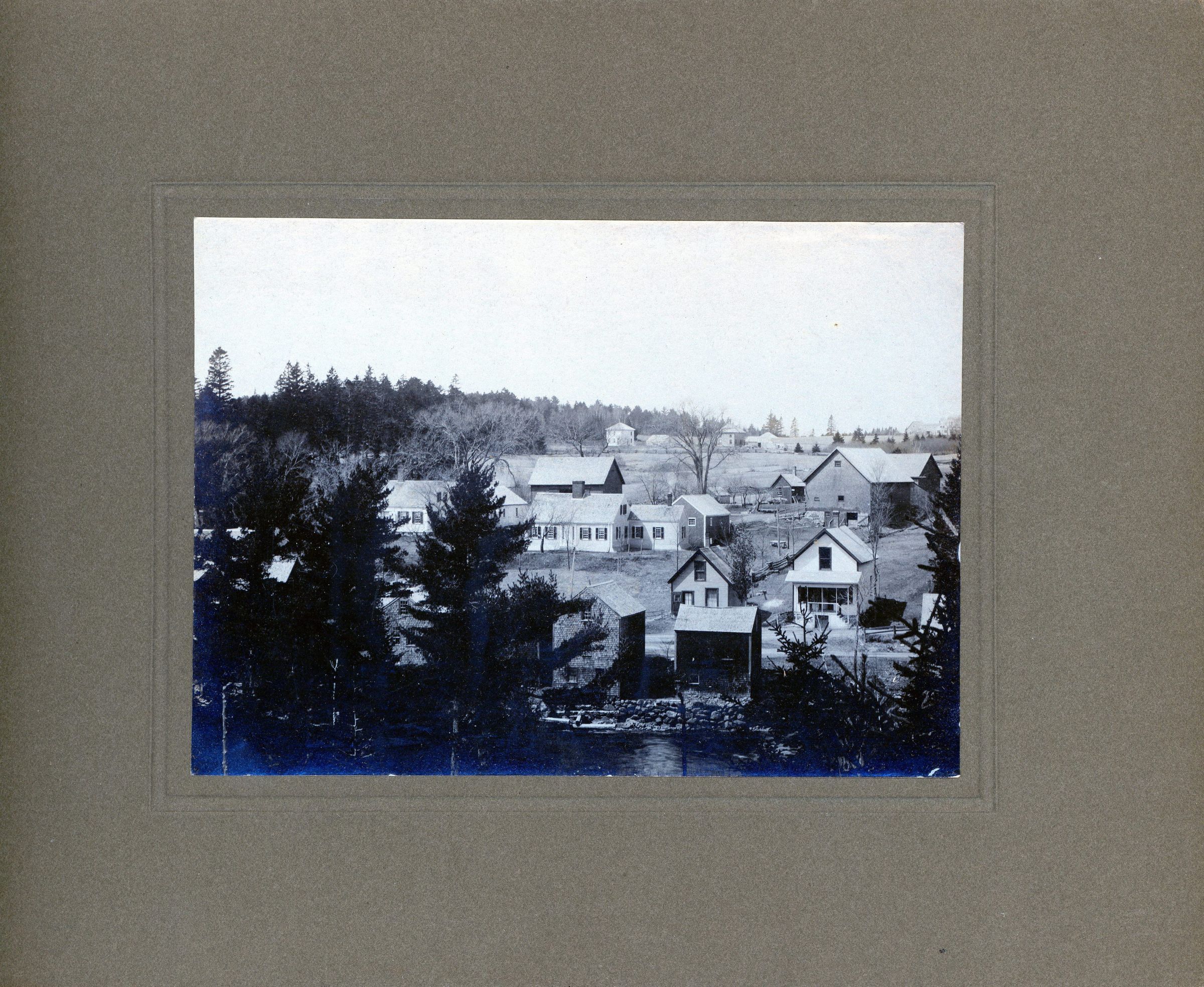 View of Dennysville, Maine from Edmunds; Four shops and storage buildings back onto the river; facing the street are two drug stores; on the hill, to the left, are Mrs. A.L.R. Gardner's house, and in the center is Fred Gardner's; across the fields on The Lane are several square Dennysville Lumber Company houses, later Archie Matheson's and Edward and Alice Burns'.