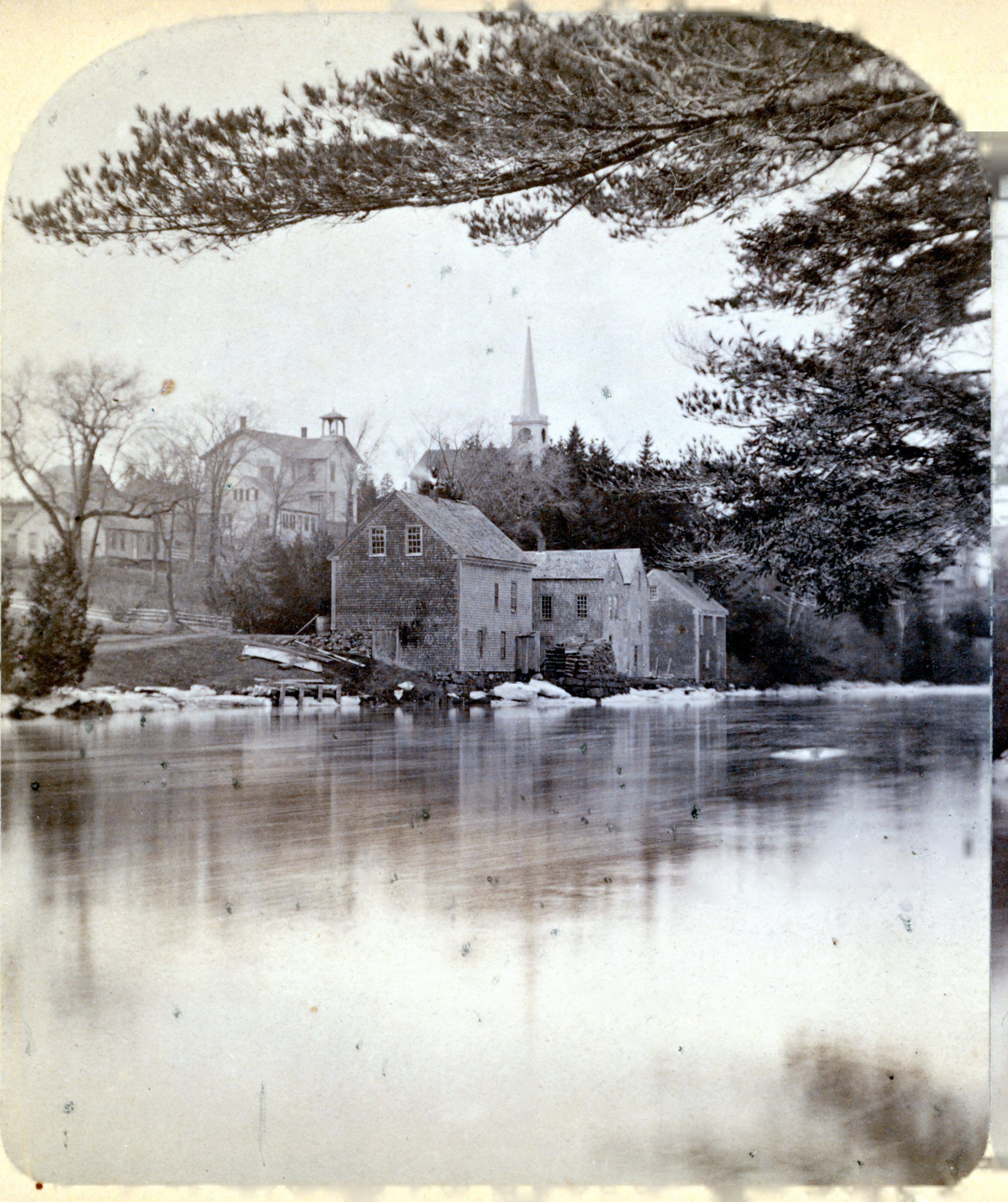Stores, Houses and School House on Meetinghouse Hill, Dennysville Maine. c. 1900; Gardner's stores line the bank o f the Dennys River, as houses across the road lead up the hill towards the Town School and Congregational Meetinghouse. William L. Kilby's house, later owned by Fred Gardner, is across from the stores, while his father William Kilby Jr.'s house is just uphill, in line with schoolhouse above, recognizable for its cupola and bell.